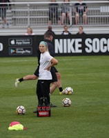 A person in sports attire stands on a soccer field, surrounded by several soccer balls and a carrier with bottles. The field is bordered by advertisements for Nike Soccer and app stores. Several people sit in bleachers in the background.