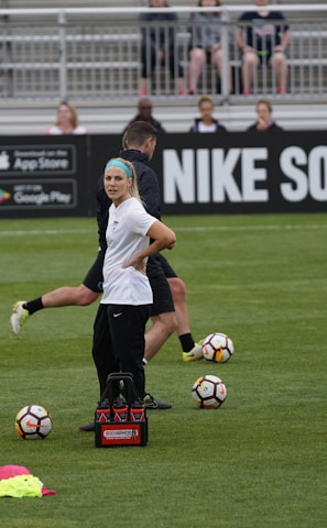 A person in sports attire stands on a soccer field, surrounded by several soccer balls and a carrier with bottles. The field is bordered by advertisements for Nike Soccer and app stores. Several people sit in bleachers in the background.