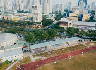 An aerial view of an urban area featuring a sports complex with a track field and a grandstand. Surrounding the sports area are numerous high-rise buildings, greenery, and roads with vehicles. The image captures people engaging in activities on the field.