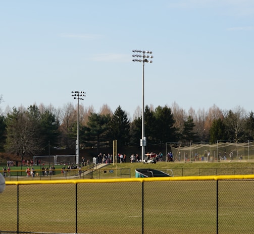 A sports field with a group of people engaging in athletic activities in the background. There is sports equipment like a soccer goal and a scoreboard visible. A chain-link fence runs across the foreground, and a statue or sculpture resembling an eagle is also seen. Trees and tall floodlights are positioned behind the field.