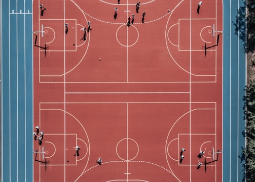 An aerial view of a large, vibrant sports complex featuring a red basketball court with multiple hoops and a surrounding blue running track. Several people are scattered across the area, engaged in various activities.