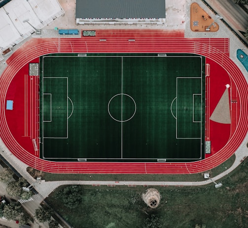 An aerial view of a large sports complex featuring a central green football field surrounded by a red running track. Adjacent to the track, there is a parking area, buildings, and pathways. Nearby, there are additional sports courts in different colors, including a blue swimming pool and orange basketball courts.
