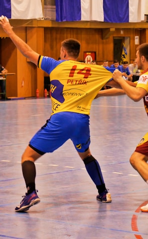 A dynamic indoor sports scene with players in motion on a court. Two athletes in blue and yellow uniforms are engaged in an intense handball game, with one player gripping the other's jersey. The background shows other players, several seated people, and advertisements on the walls.