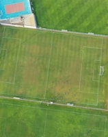 An aerial view of a sports complex featuring multiple green grass fields, including football pitches with marked goalposts. Adjacent to the fields is a blue-roofed building and a smaller area with a tennis or basketball court. Surrounding the complex, there's a road lined with trees and a dirt area with sparse vegetation.