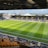 A sports stadium with a well-maintained grass field is depicted. Players are spread across the field, engaging in warm-up activities. The stands are partially filled with spectators, and the stadium is surrounded by urban buildings under a partly cloudy sky.