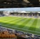 A sports stadium with a well-maintained grass field is depicted. Players are spread across the field, engaging in warm-up activities. The stands are partially filled with spectators, and the stadium is surrounded by urban buildings under a partly cloudy sky.