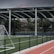 An outdoor sports facility featuring a running track and a soccer goal. The track is red, surrounded by a black metal fence, and the field is covered with artificial green turf. In the background, a basketball court is visible under a covered roof with metal supports. The sky is overcast, adding a moody atmosphere.