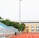 A sports stadium with a red running track, blue seating area, and a large floodlight. The background features a yellow building with blue and white accents, surrounded by green trees. The atmosphere is clean and empty, with clear skies above.