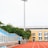 A sports stadium with a red running track, blue seating area, and a large floodlight. The background features a yellow building with blue and white accents, surrounded by green trees. The atmosphere is clean and empty, with clear skies above.