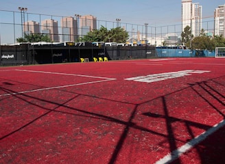 A sports court with a red playing surface and white lines, surrounded by a black mesh fence. In the background, tall buildings are visible, along with some trees and a few yellow chairs positioned near the fence.