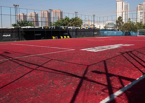 A sports court with a red playing surface and white lines, surrounded by a black mesh fence. In the background, tall buildings are visible, along with some trees and a few yellow chairs positioned near the fence.
