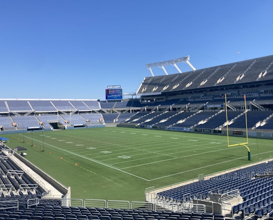 A large, empty sports stadium with blue seats surrounding a well-maintained green grass field. Bright sunlight creates clear shadows, and there are two goal posts on opposite ends of the field. A big electronic scoreboard displays some graphics, and the sky is clear and blue.